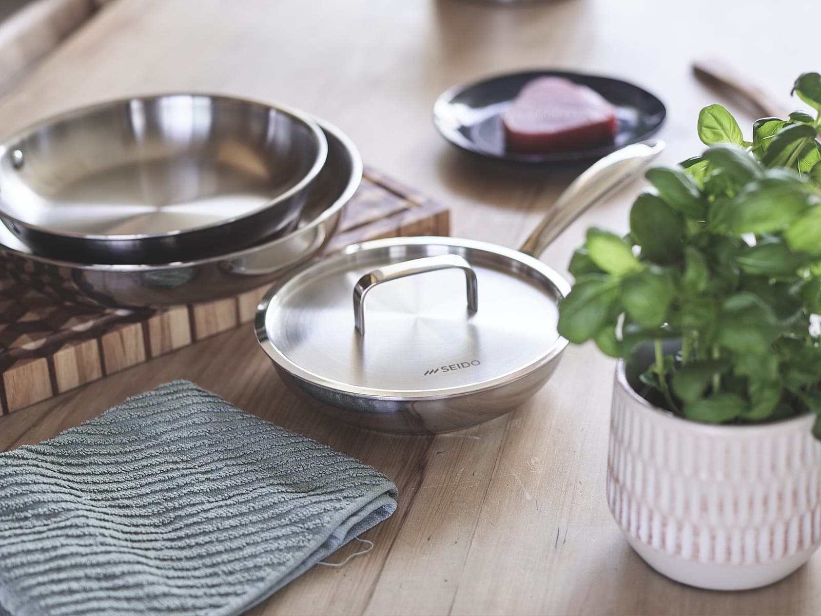 Stainless steel skillets with their lid on a kitchen table beside steak and basil